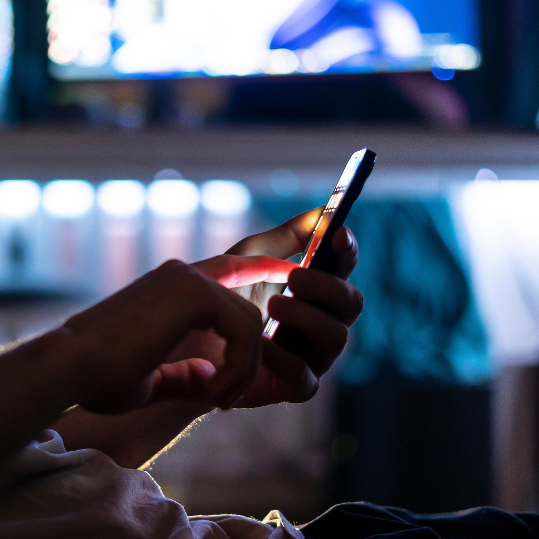 Hands using a smartphone with a blurred TV in the background, illustrating competing screens for creative advertising.